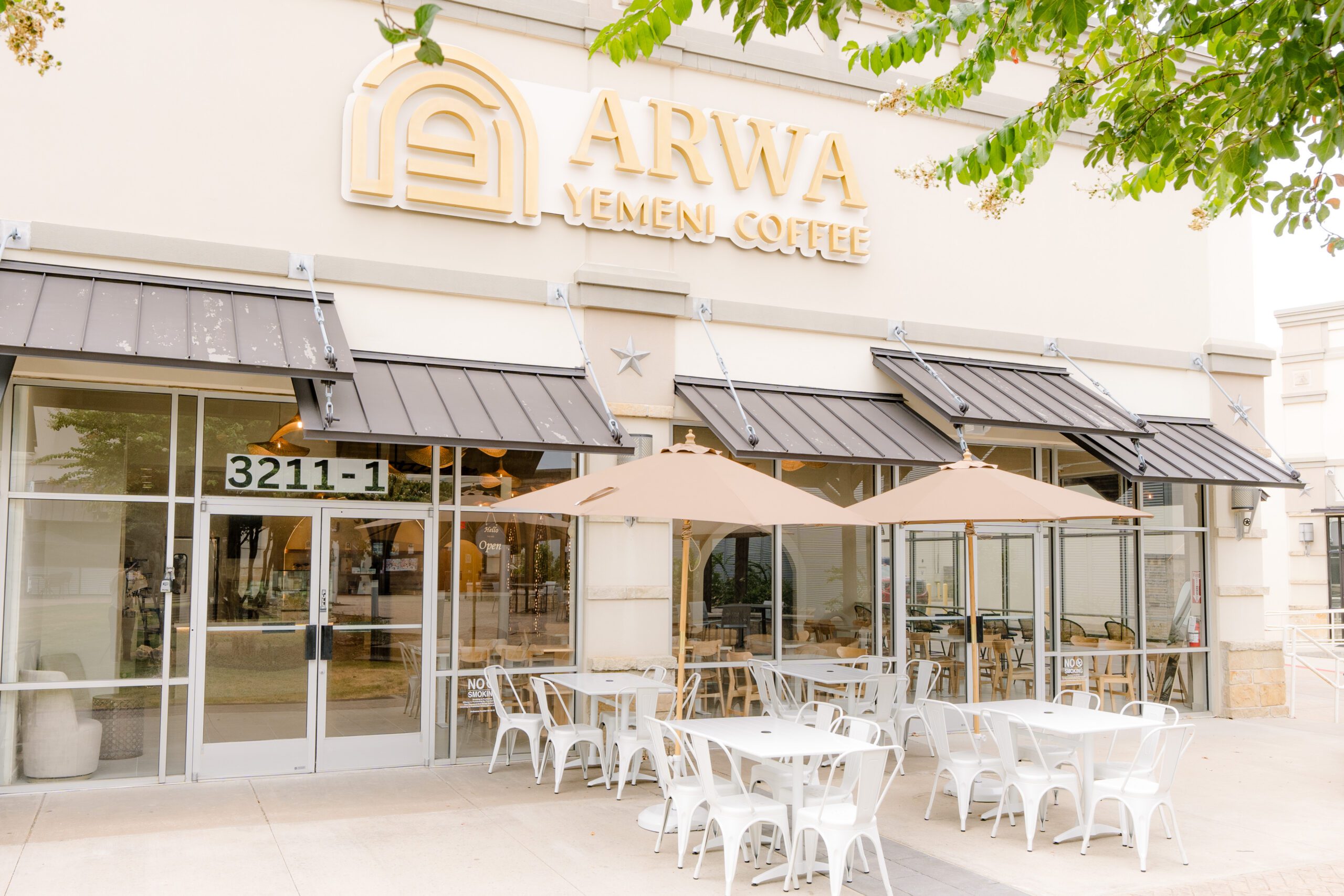 An Arwa Yemeni Coffee storefront — cream stucco, dark bronze awnings, white outdoor furniture beneath beige umbrellas, the ARWA wordmark with the arched logo above the door.
